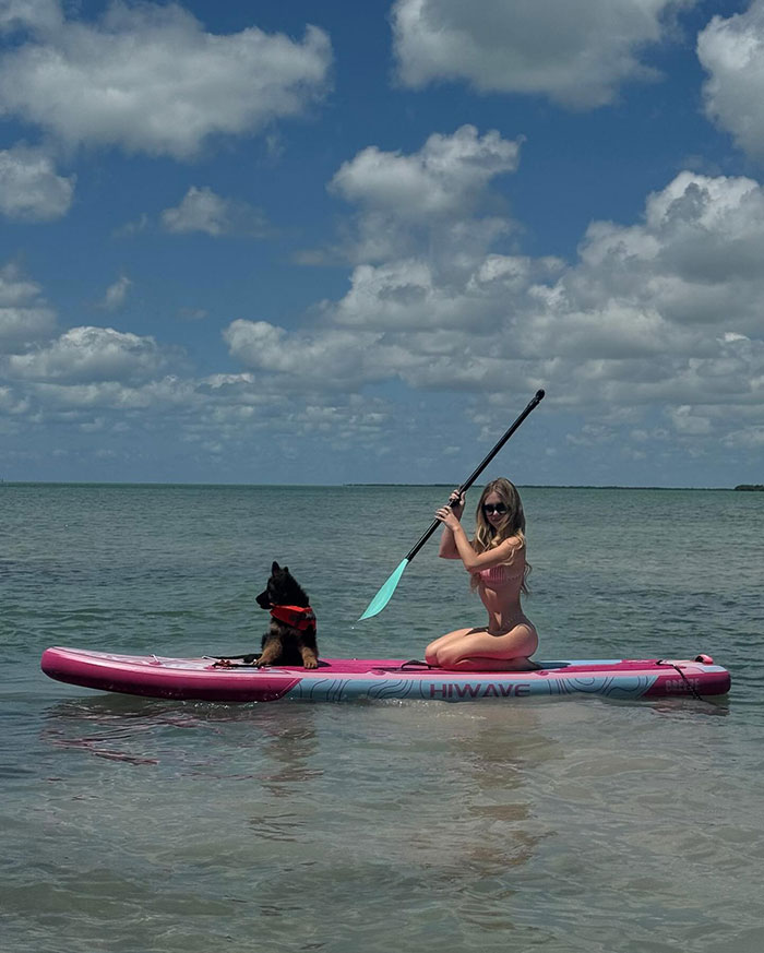 Sydney Sweeney on paddleboard with puppy in red harness, on calm water under partly cloudy sky, enjoying outdoor activity. Sydney Sweeney on paddleboard with puppy in red harness, on calm water under partly cloudy sky, enjoying outdoor activity.