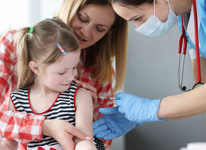 Young girl receiving vaccine from masked healthcare worker as mother looks on in medical setting related to anti-vaxxer consequences. Young girl receiving vaccine from masked healthcare worker as mother looks on in medical setting related to anti-vaxxer consequences.