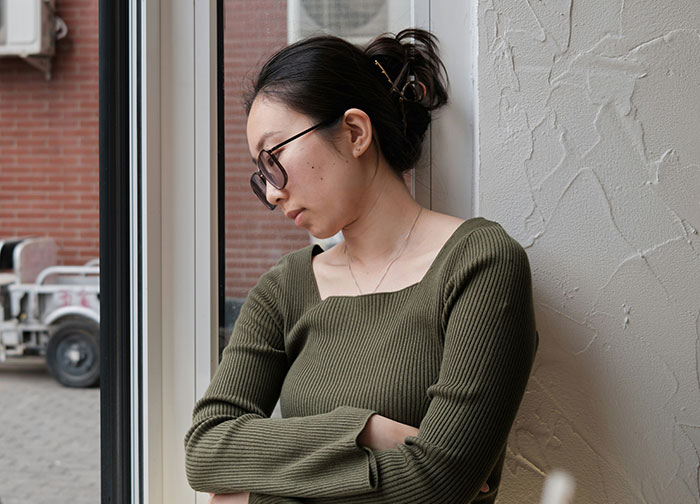 Woman wearing sunglasses and green ribbed sweater standing by window with arms crossed, reflecting on neighbor complaints about outfit. Woman wearing sunglasses and green ribbed sweater standing by window with arms crossed, reflecting on neighbor complaints about outfit.