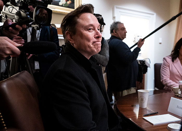 Elon Musk sitting at a table during a media event as Tesla searches for new CEO to replace him. Elon Musk sitting at a table during a media event as Tesla searches for new CEO to replace him.