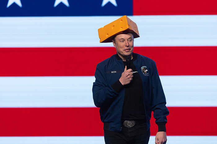 Man speaking on stage with microphone, wearing a cheese hat and jacket, against an American flag backdrop, Tesla CEO search topic. Man speaking on stage with microphone, wearing a cheese hat and jacket, against an American flag backdrop, Tesla CEO search topic.