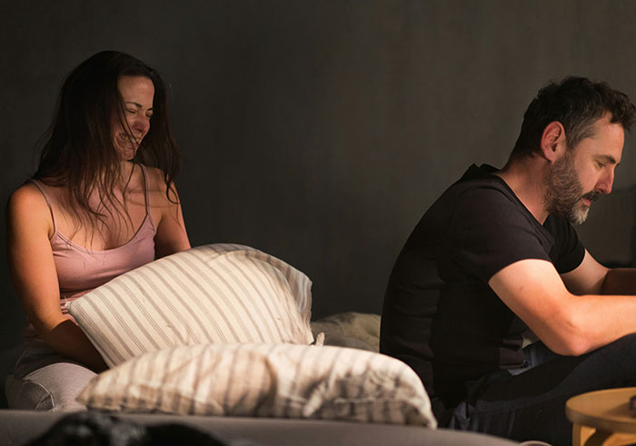 A woman holding a pillow and a stressed man sitting on a bed depicting moments that could destroy people’s lives.