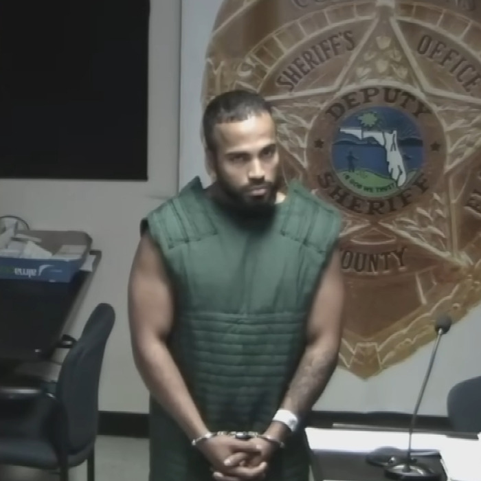 Man in green detention vest and handcuffs standing in front of sheriff's office emblem during a court appearance. Man in green detention vest and handcuffs standing in front of sheriff's office emblem during a court appearance.