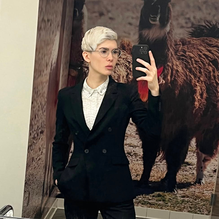 Trans archer with short blonde hair and glasses takes a mirror selfie wearing a black suit jacket in a room with animal wall art. Trans archer with short blonde hair and glasses takes a mirror selfie wearing a black suit jacket in a room with animal wall art.