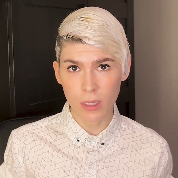 Trans archer with short blonde hair wearing a patterned shirt, speaking in an indoor setting with a serious expression. Trans archer with short blonde hair wearing a patterned shirt, speaking in an indoor setting with a serious expression.