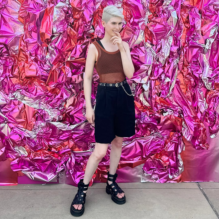 Young trans archer standing against vibrant pink metallic background, dressed in casual black shorts and a mesh top. Young trans archer standing against vibrant pink metallic background, dressed in casual black shorts and a mesh top.
