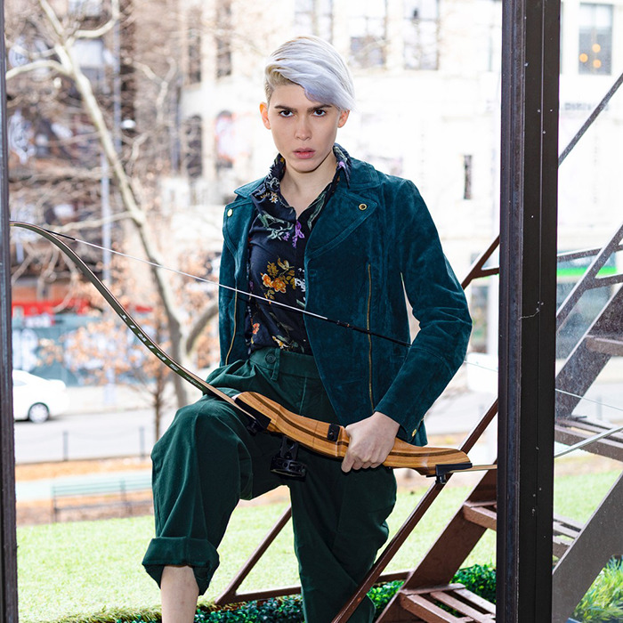 Trans archer with silver hair holding a bow outdoors, wearing a teal jacket and floral shirt, posing confidently near stairs. Trans archer with silver hair holding a bow outdoors, wearing a teal jacket and floral shirt, posing confidently near stairs.