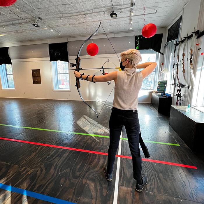Trans archer practicing indoors aiming bow, wearing mask and light sleeveless top, with bows displayed on wall. Trans archer practicing indoors aiming bow, wearing mask and light sleeveless top, with bows displayed on wall.