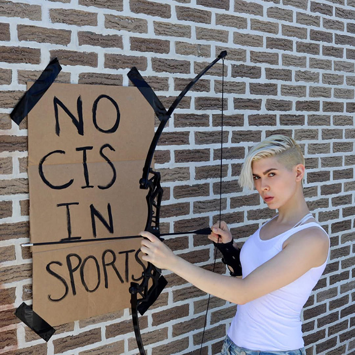 Trans archer aiming bow near wall with no cis in sports sign, illustrating backlash faced in competitive sports context. Trans archer aiming bow near wall with no cis in sports sign, illustrating backlash faced in competitive sports context.