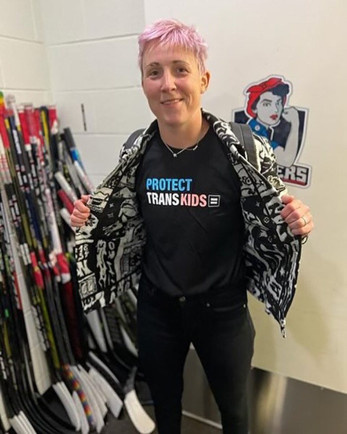 Female athlete showing support for trans kids wearing jacket open in a hockey locker room with sticks in background Female athlete showing support for trans kids wearing jacket open in a hockey locker room with sticks in background