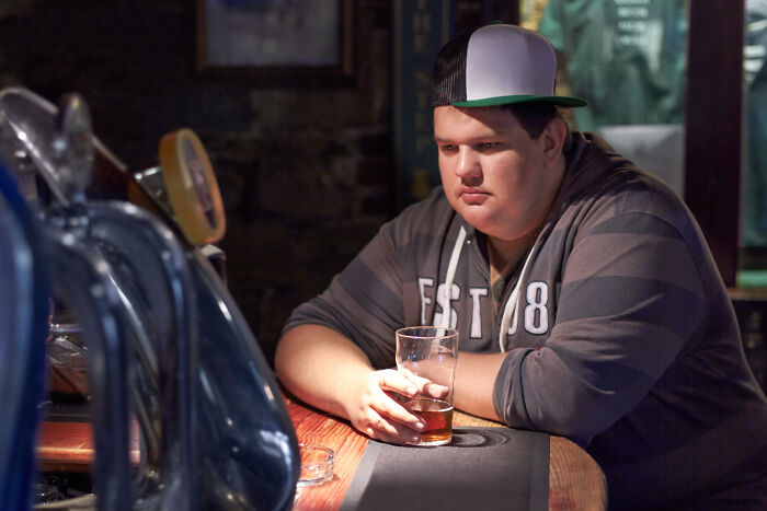 Young man appearing unhealthy and reflective, sitting alone at a bar holding a glass, illustrating unhealthy human things.