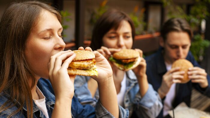 Three young people eating burgers outdoors, showing unhealthy human things in their casual meal choices.