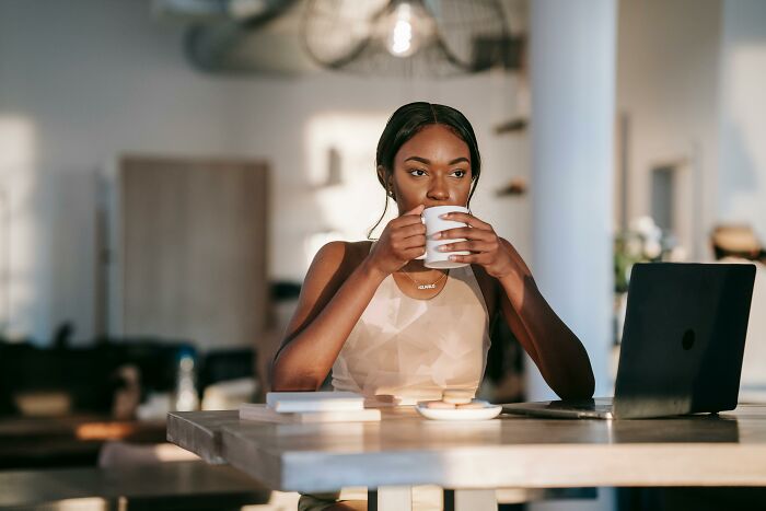Young woman drinking from a cup while sitting at a table with laptop and books, highlighting unhealthy human things.