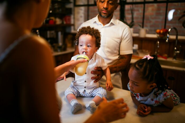 A family in a kitchen with a toddler drinking from a bottle, highlighting unhealthy human things in everyday life.