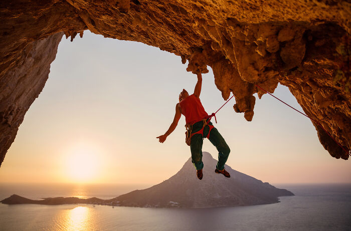 Person climbing a rocky cave over water at sunset, illustrating unhealthy human things like risky physical activities.