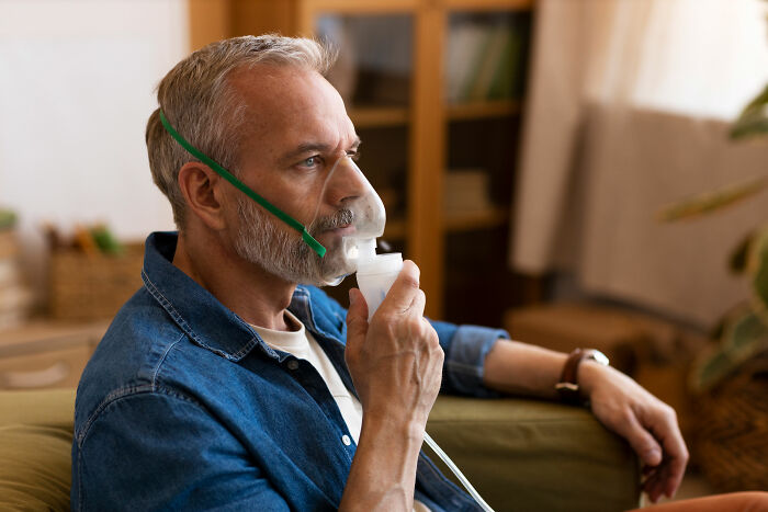 Middle-aged man using oxygen mask at home, highlighting unhealthy human things related to respiratory issues and health.