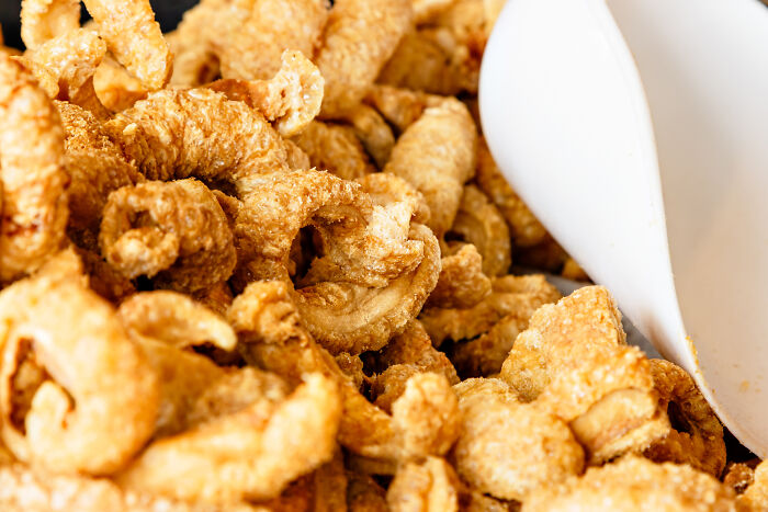 Close-up of unhealthy human things pork rinds with a white scoop resting on crispy chips in a bowl.
