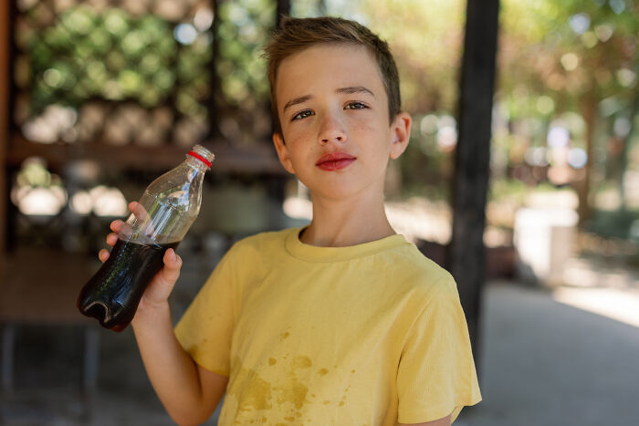 Young boy wearing a yellow shirt with stains, holding a soda bottle, illustrating unhealthy human things related to diet choices outdoors.