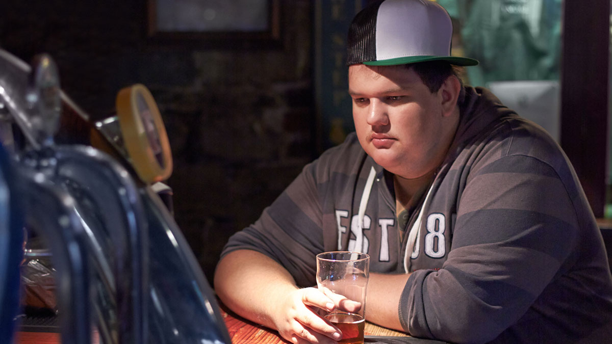 Overweight young man wearing a cap sitting alone at a bar with a drink, reflecting on unhealthy human things.
