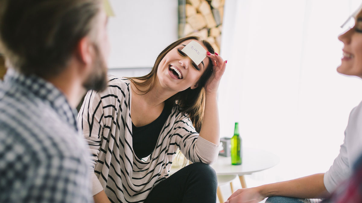 Young woman laughing with sticky note on forehead playing a game, showing hilarious main character moments with friends.