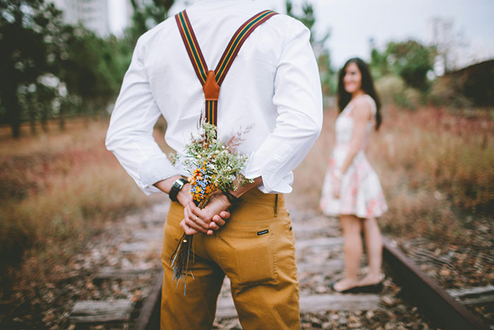 Man holding flowers behind his back wearing suspenders, woman standing on railway tracks in floral dress, wedding theme. Man holding flowers behind his back wearing suspenders, woman standing on railway tracks in floral dress, wedding theme.