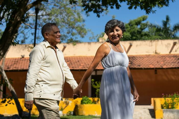 Elderly couple holding hands outdoors in bright sunlight near a house with a tiled roof and garden.