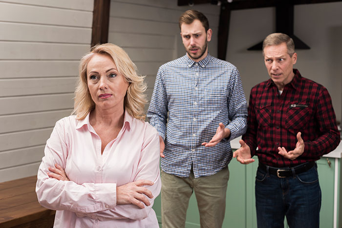 Woman standing with arms crossed while two men talk to her, illustrating dog mom DIL demanding equal treatment after Mother’s Day gifting. Woman standing with arms crossed while two men talk to her, illustrating dog mom DIL demanding equal treatment after Mother’s Day gifting.