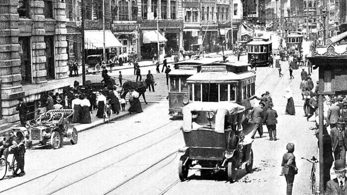 Busy street scene from 100 years ago in America showing vintage cars, streetcars, and people dressed in early 20th century attire.