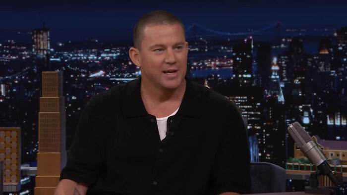 Man in black shirt speaking on late-night talk show set against city skyline backdrop, discussing mind-boggling beliefs.