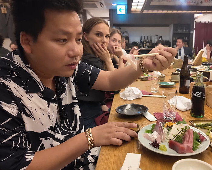 Vlogger sitting at a restaurant table with sushi, recording a video unaware of the A-list star nearby. Vlogger sitting at a restaurant table with sushi, recording a video unaware of the A-list star nearby.