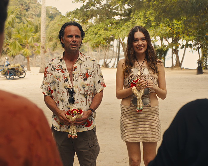 Walton Goggins and Aimee Lou Wood holding floral garlands outdoors in a tropical setting during a casual moment. Walton Goggins and Aimee Lou Wood holding floral garlands outdoors in a tropical setting during a casual moment.