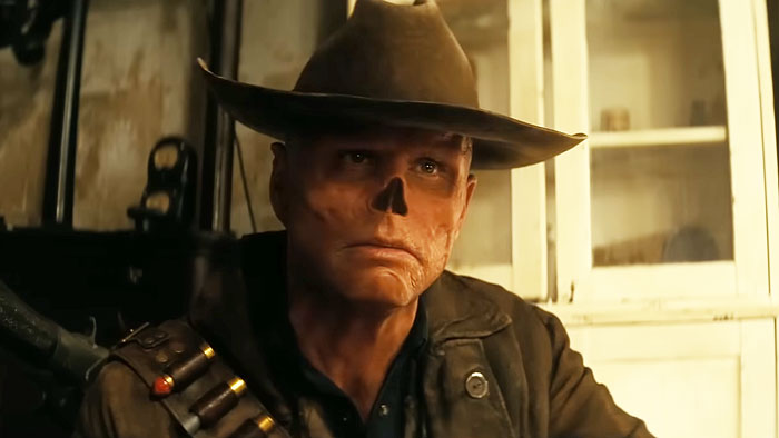 Walton Goggins wearing a wide-brimmed hat and dark jacket in a dimly lit setting portraying a rugged character.