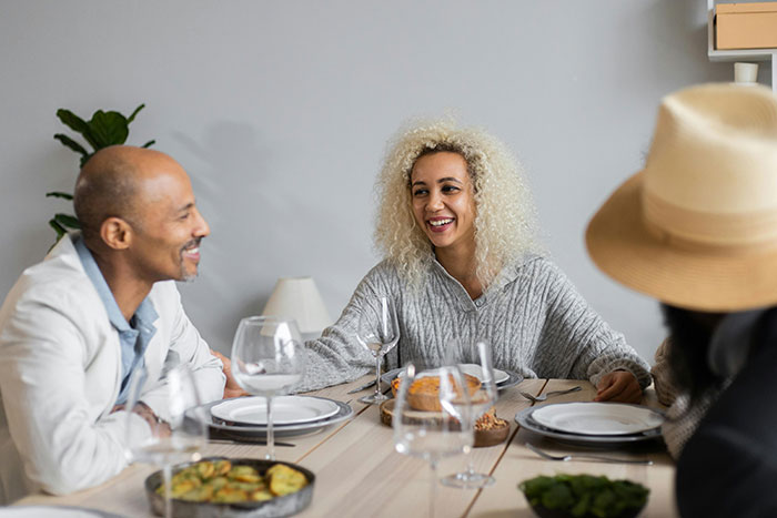 Man smiling at woman with curly hair at dining table with wife’s best friend, capturing a tense social moment. Man smiling at woman with curly hair at dining table with wife’s best friend, capturing a tense social moment.