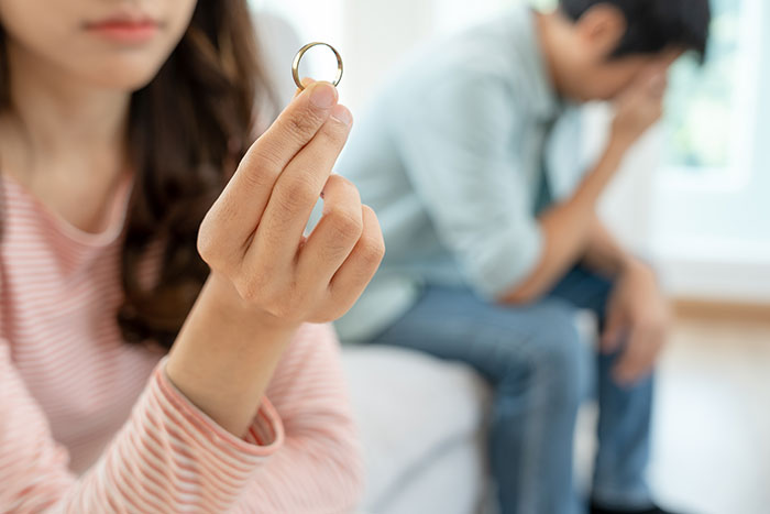 Wife holding wedding ring looking upset, husband sitting in background with head in hands, emotional conflict visible. Wife holding wedding ring looking upset, husband sitting in background with head in hands, emotional conflict visible.