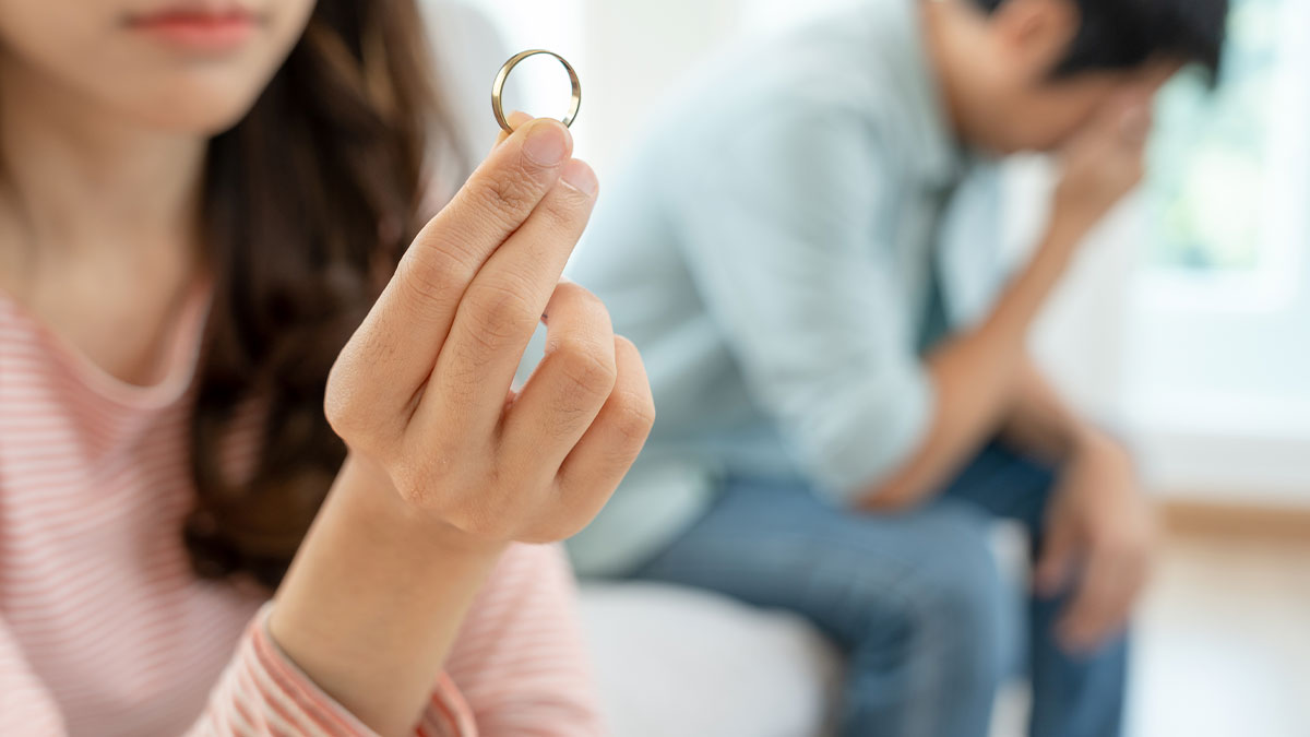 Wife holding wedding ring sadly while husband sits behind emotionally checked out in a tense moment.