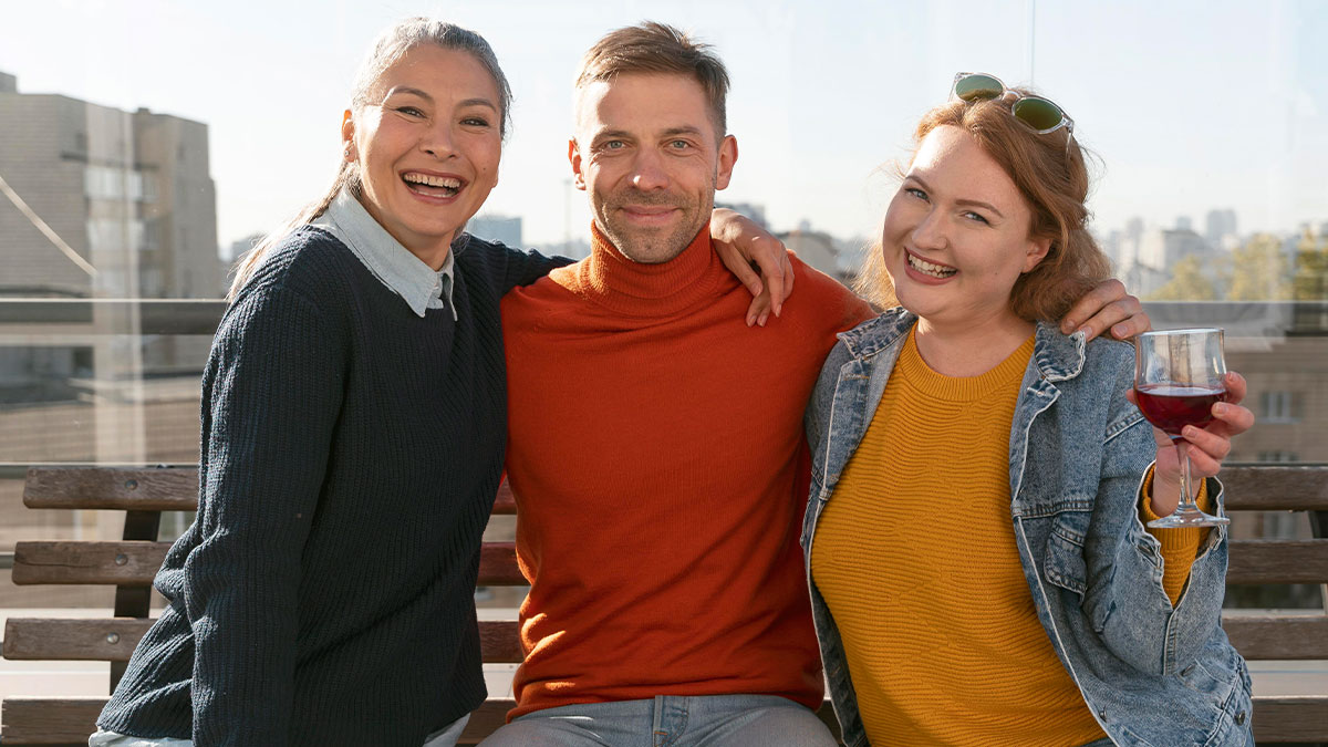 Three friends sitting closely on a bench outdoors, smiling and enjoying a casual catch-up with drinks.