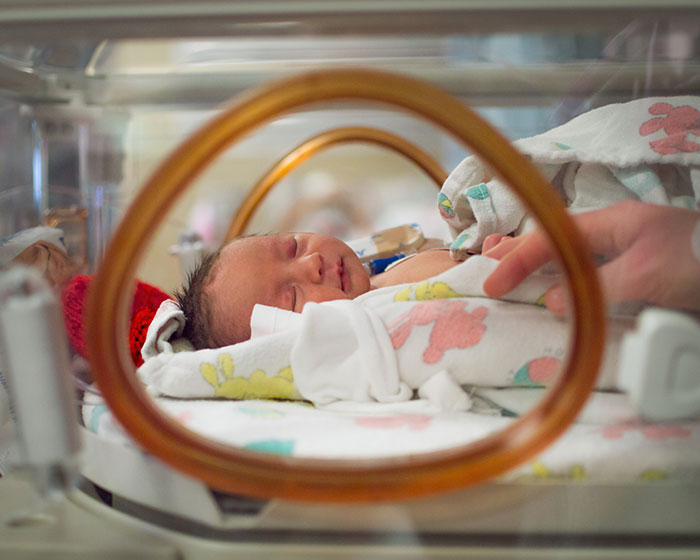 Newborn baby resting in a hospital incubator with a hand gently touching the blanket, highlighting family and care. Newborn baby resting in a hospital incubator with a hand gently touching the blanket, highlighting family and care.