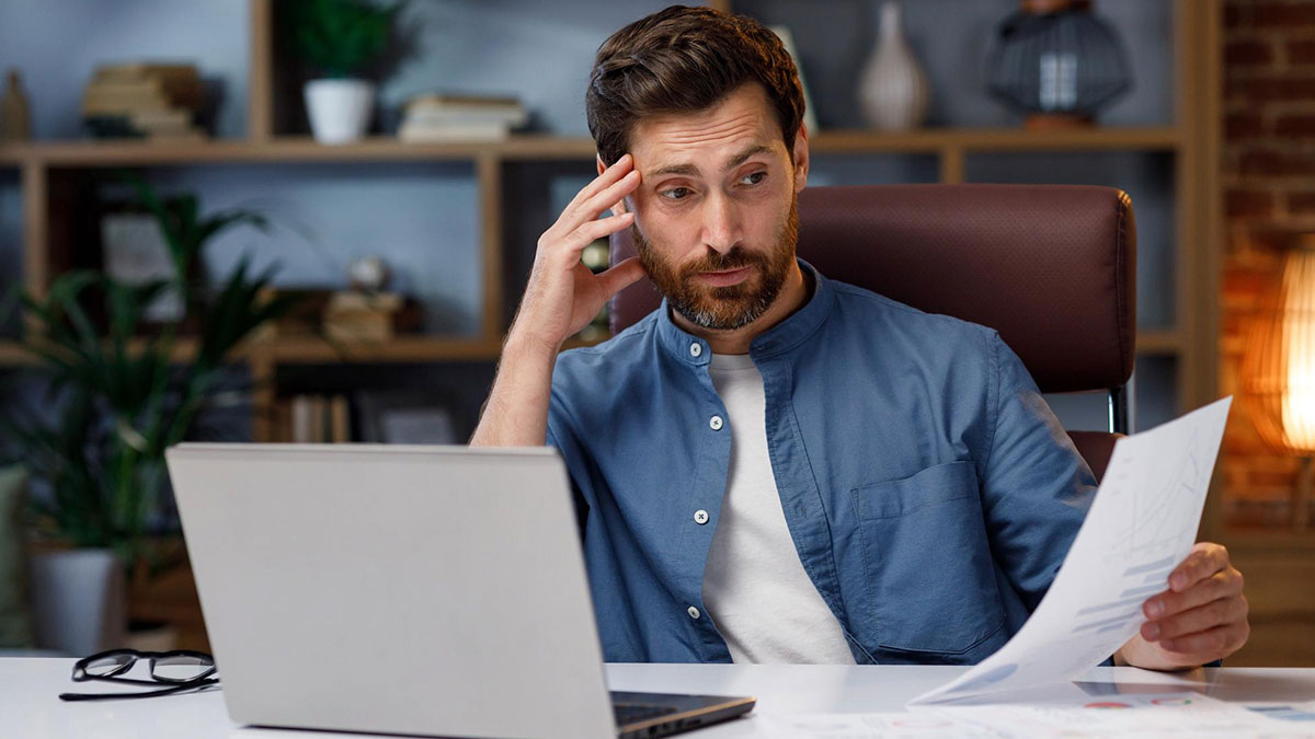 Man sitting at desk looking stressed while reviewing papers and laptop, representing wildest life blunders and secrets.