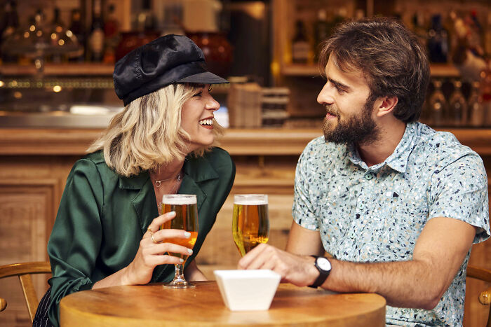 A smiling couple enjoying beers at a cozy bar, sharing a moment reflecting on life-changing small decisions.