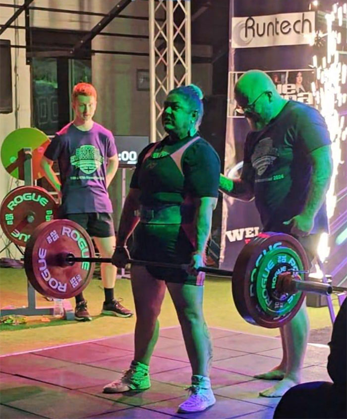 Woman powerlifting in competition with coaches nearby, lifting a heavy barbell amid event lighting and spectators. Woman powerlifting in competition with coaches nearby, lifting a heavy barbell amid event lighting and spectators.