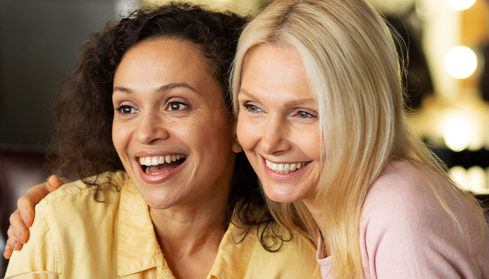 Two women smiling together, representing a woman setting boundaries with her friend about hosting a toddler. Two women smiling together, representing a woman setting boundaries with her friend about hosting a toddler.