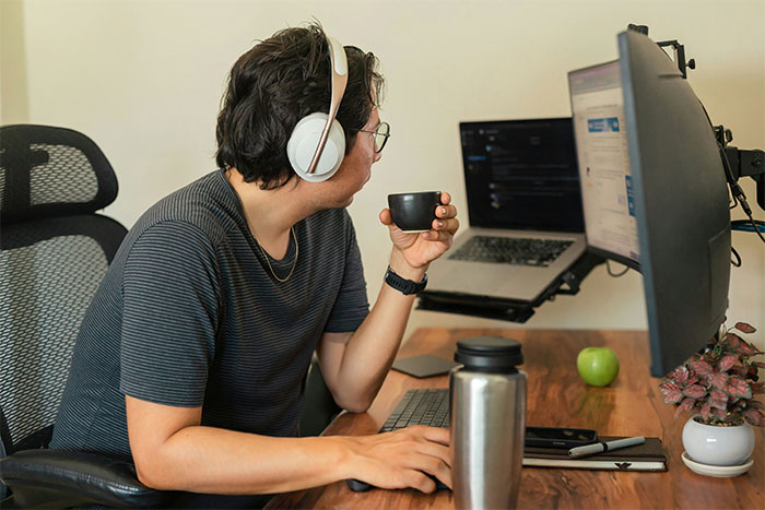 Employee following unworkable deployment rule wearing headphones, working at dual monitors, and holding a cup in office setting. Employee following unworkable deployment rule wearing headphones, working at dual monitors, and holding a cup in office setting.