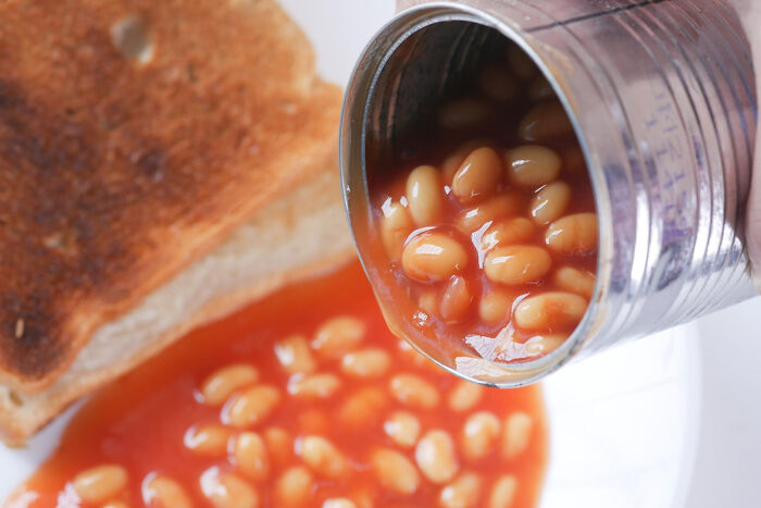 Can of processed baked beans pouring onto a plate with toast, illustrating best processed foods for weight loss. Can of processed baked beans pouring onto a plate with toast, illustrating best processed foods for weight loss.