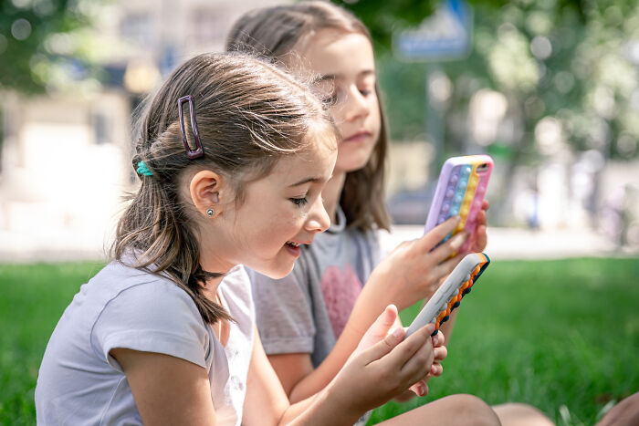 Two young girls sitting on grass outdoors, smiling and focused on their phones, showing a good trend slowly disappeared.