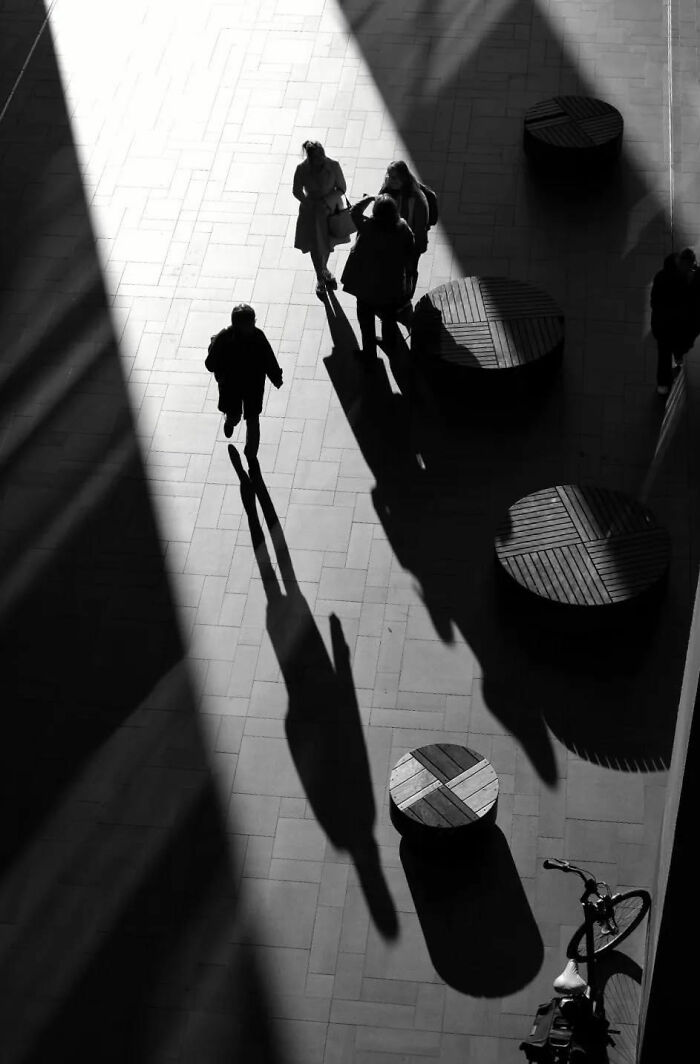 Black and white street shot capturing long shadows of people walking and wooden benches in urban everyday life scene.