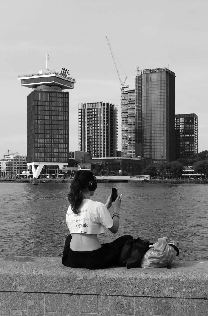 Person with headphones sitting by river, capturing street shots of city buildings in everyday life scene.
