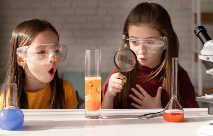 Two girls wearing safety goggles, observing a chemical reaction with curiosity, showcasing a good trend slowly disappeared in science learning.
