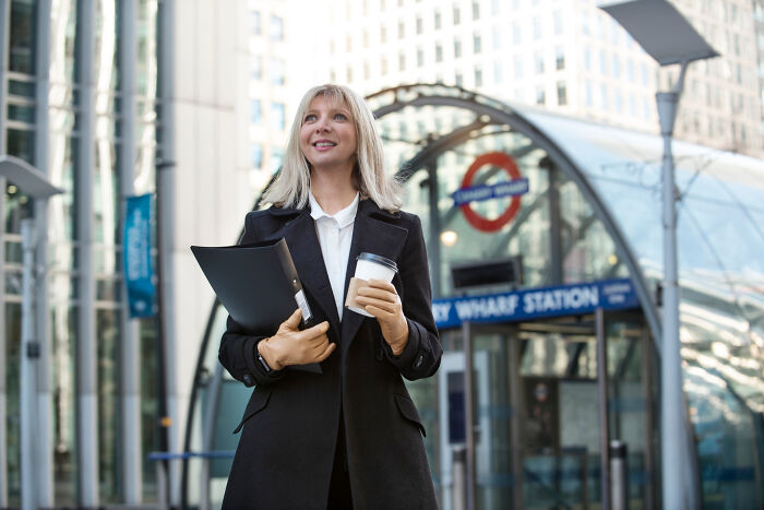 Professional woman holding coffee and documents outside Canary Wharf Station, reflecting honest thoughts about the UK.