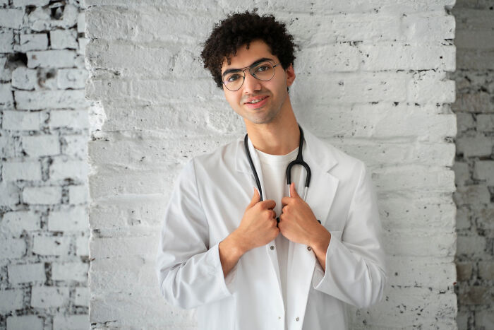 Young man dressed as a doctor wearing glasses and a white coat, pretending medical expertise against a brick wall background.