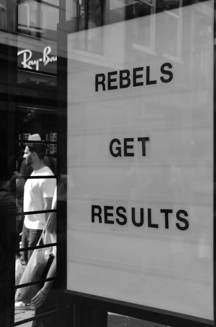 Black and white street shot showing a sign reading rebels get results with people walking by, capturing the poetry of everyday life.